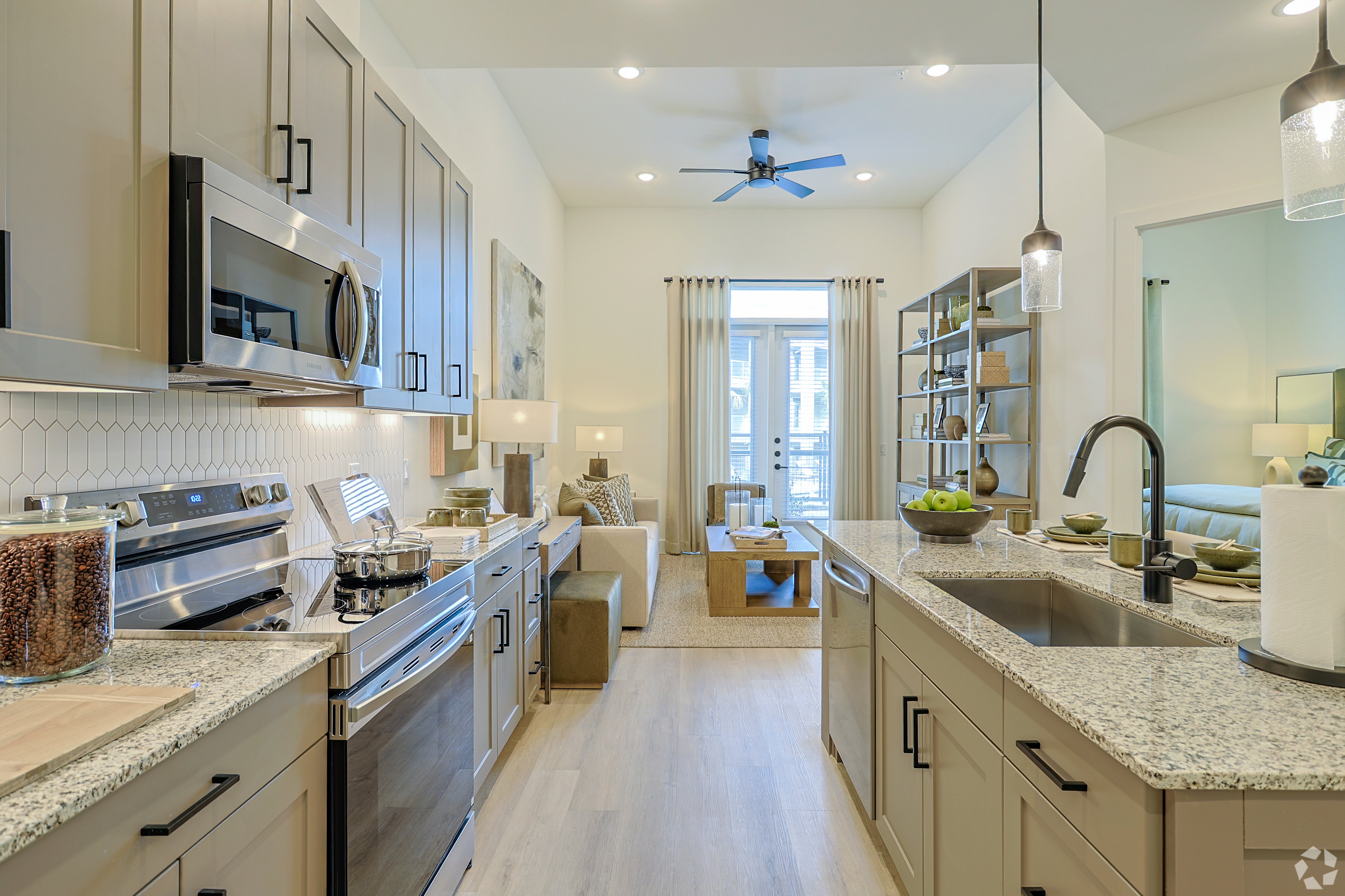 A modern kitchen with stainless steel appliances and granite countertops opens up to a bright living area with light-colored furniture and a balcony in the background.