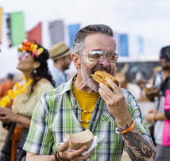 A man in a green plaid shirt is eating a corn dog at an outdoor event.