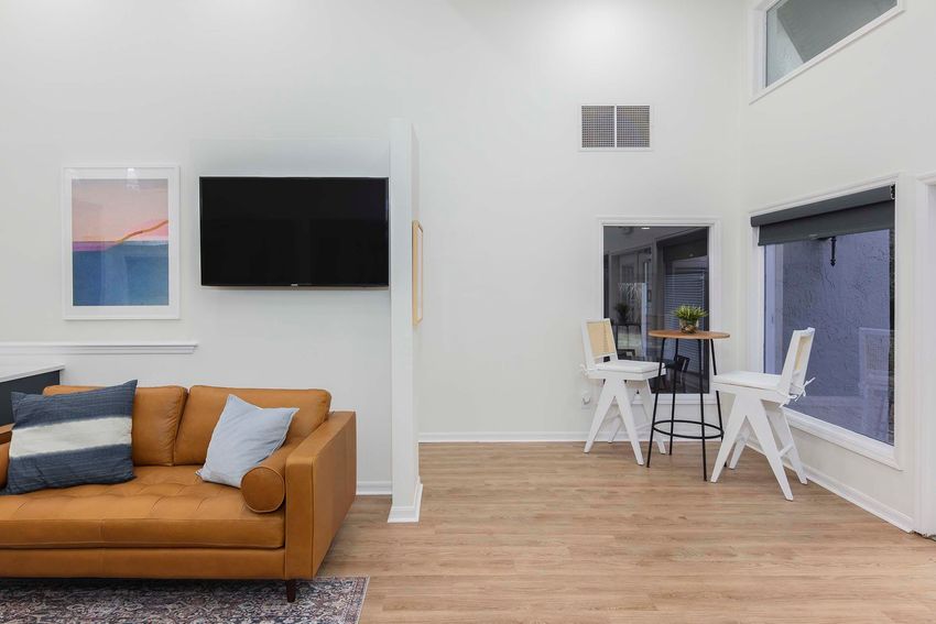 A modern living space featuring a tan leather sofa adorned with blue and white cushions. Opposite the sofa, a wall-mounted television is visible, alongside minimalist furniture including a small round table with two chairs by a window. Light wood floors and neutral walls create a bright atmosphere.