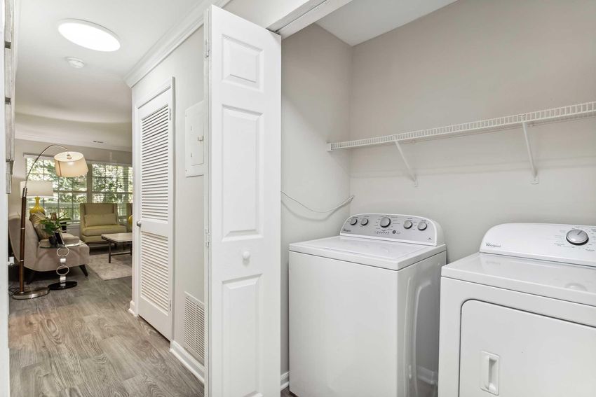 A spacious laundry room featuring a washer and dryer side by side, with a shelf above for storage. The room has light-colored walls and a doorway leading to a bright living area with natural light. A comfortable seating area can be seen in the background, creating an inviting space.
