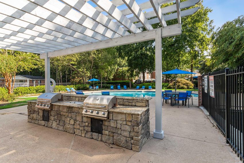 Outdoor pool area featuring a stone barbecue grill station, tables with blue umbrellas, and lounge chairs surrounding a sparkling swimming pool, all set under a wooden trellis with a clear sky and lush green trees in the background.