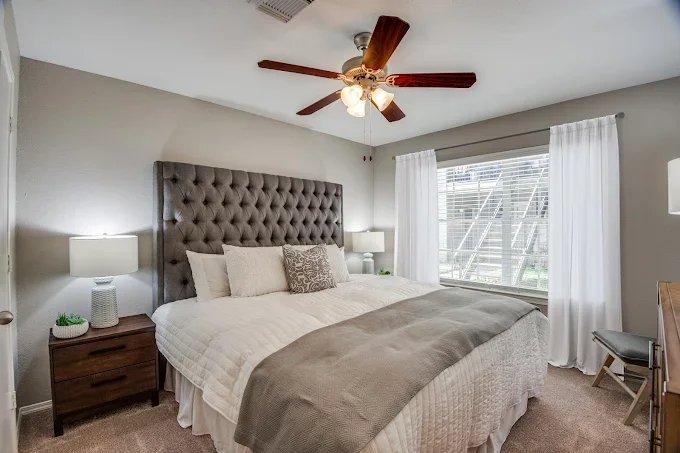 Bedroom with large bed, tufted gray headboard, white bedding, decorative pillows, two nightstands with lamps, large window with white curtains, ceiling fan with wooden blades, beige carpet, and a wooden desk with a gray chair.