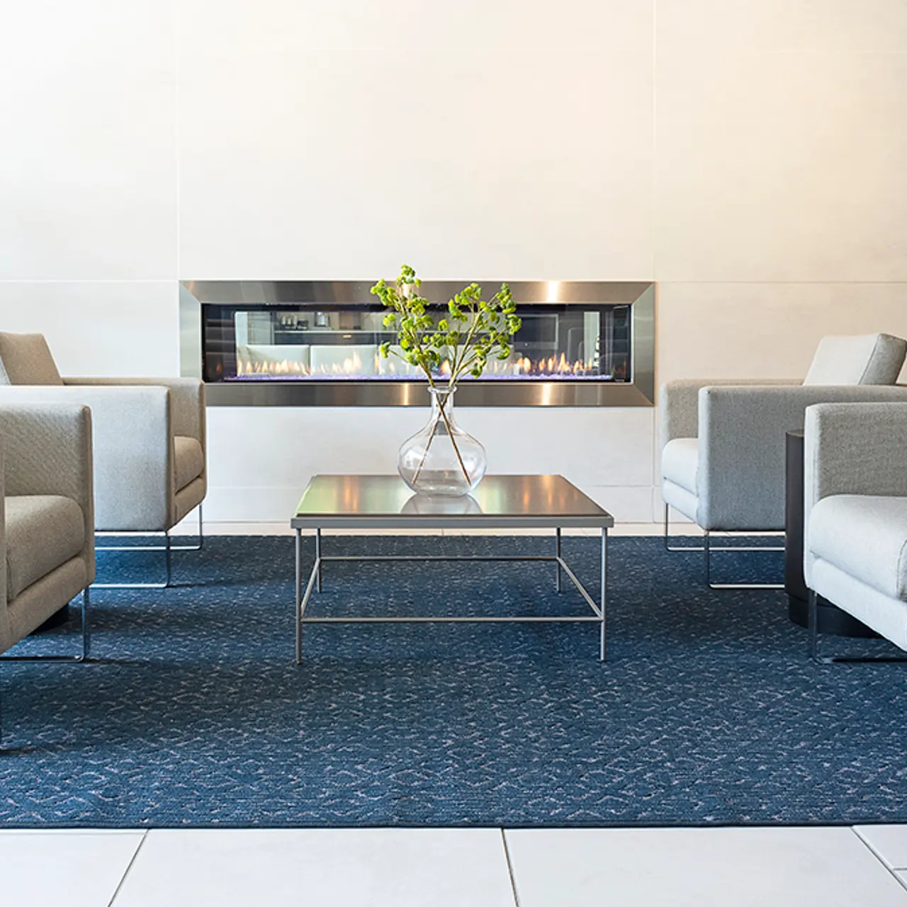 A modern lounge area featuring four light gray armchairs arranged around a glass table, with a decorative vase and greenery on the table. In the background, there's an elegant fireplace mounted on a wall. The floor has light-colored tiles and a blue patterned rug beneath the furniture.