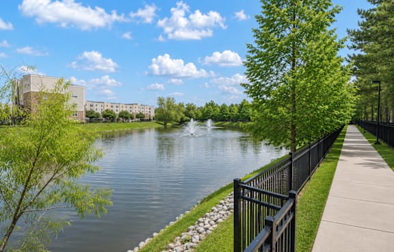 A serene lake with a fountain in the middle, surrounded by a black fence and green trees.