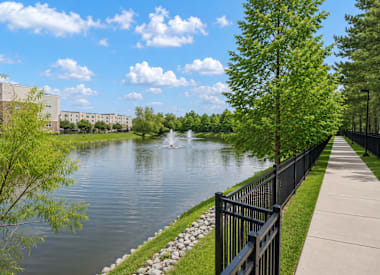 A serene lake with a fountain in the middle, surrounded by a black fence and green trees.