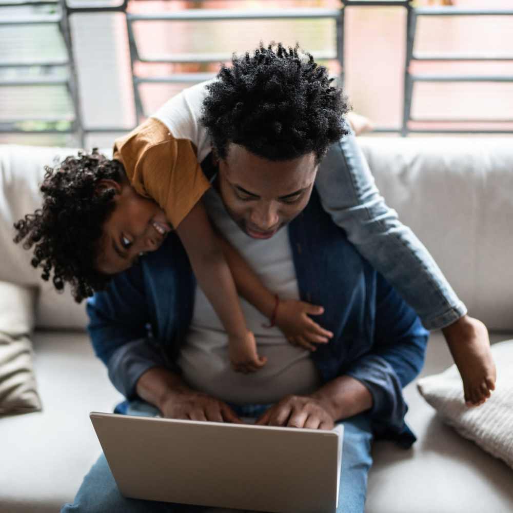 Father working on laptop with toddler playfully hanging on his back in modern living room at Riverwood Apartments in Wilmington, North Carolina