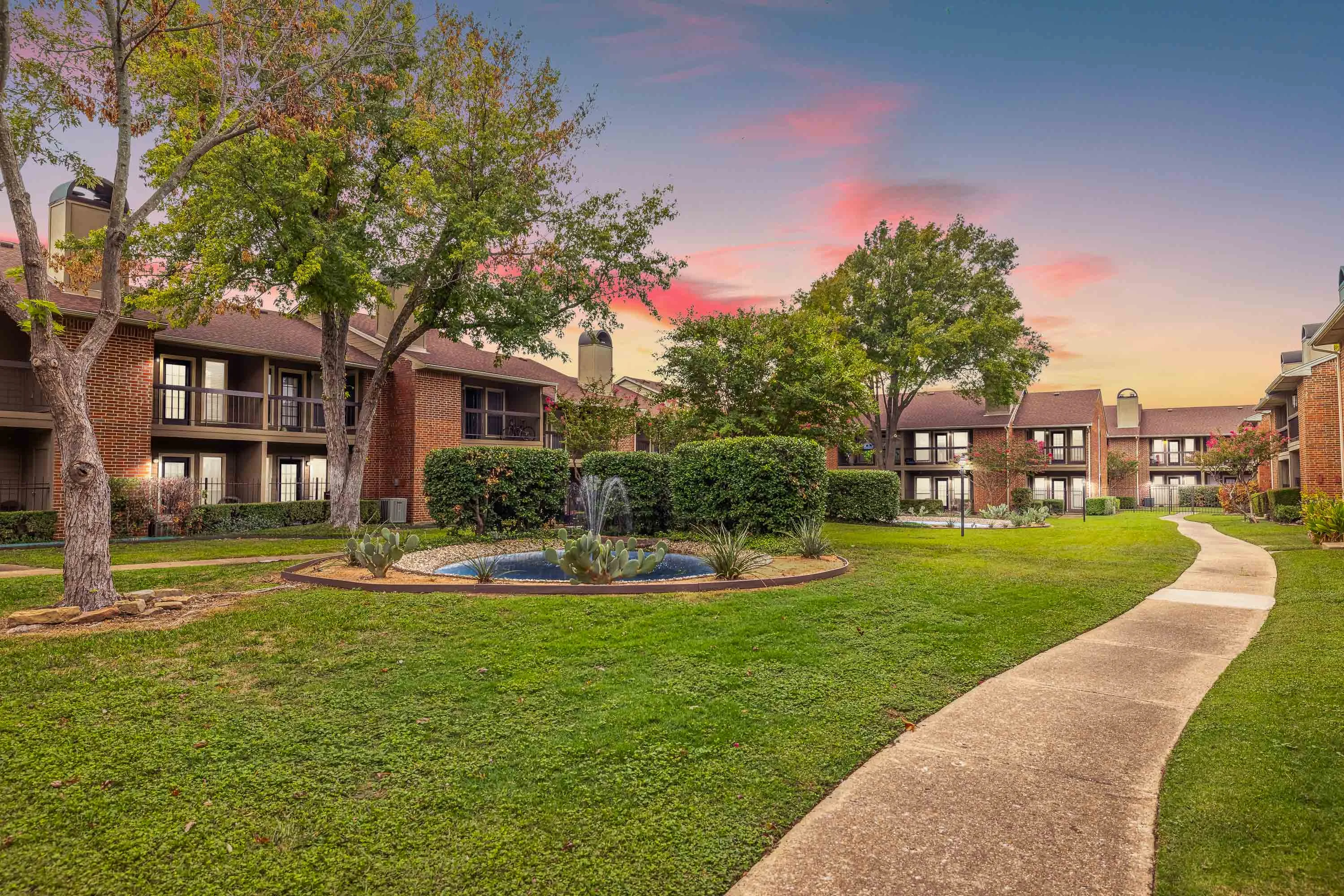 Walking path past a fountain, landscaping, and shade trees in an apartment community with two story brick apartment buildings 