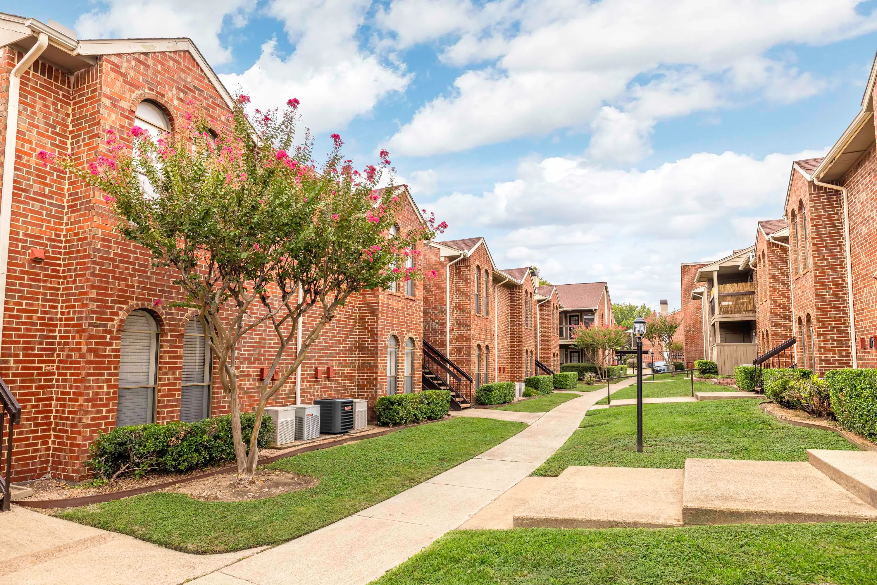 Walking path between apartment buildings with arched windows and brick exterior on a mostly cloudy day 