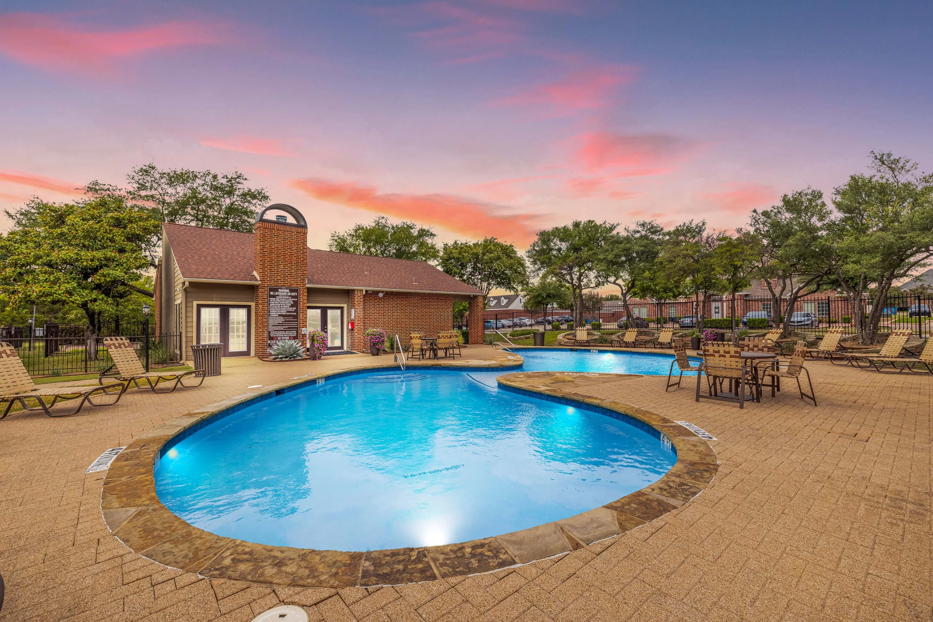 Swimming pool with a stone patio with lounge chairs and tables surrounded by a black fence at dusk 