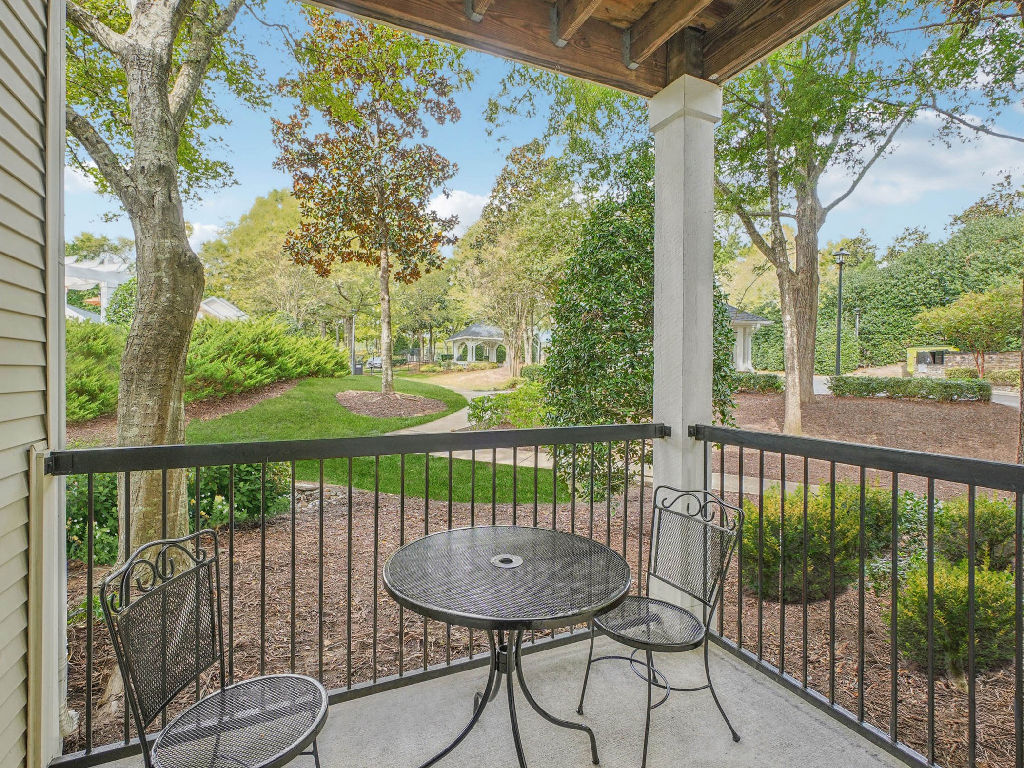 Covered patio with a round metal table and two chairs overlooks a landscaped garden with trees, bushes, and a walkway leading to a gazebo.