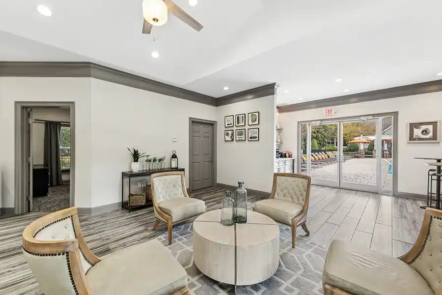 A modern lounge area with four beige chairs around a round table, neutral decor, framed art on the wall, and glass doors leading to an outdoor patio.