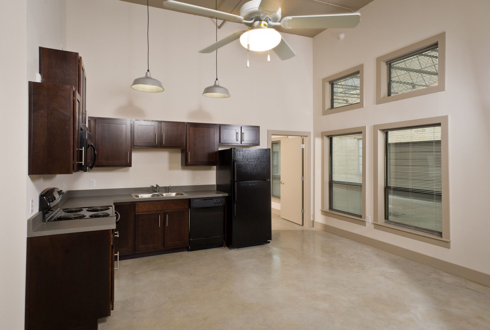 Industrial-style apartment kitchen with high, exposed ceilings, large windows, warm espresso cabinets, black appliances.