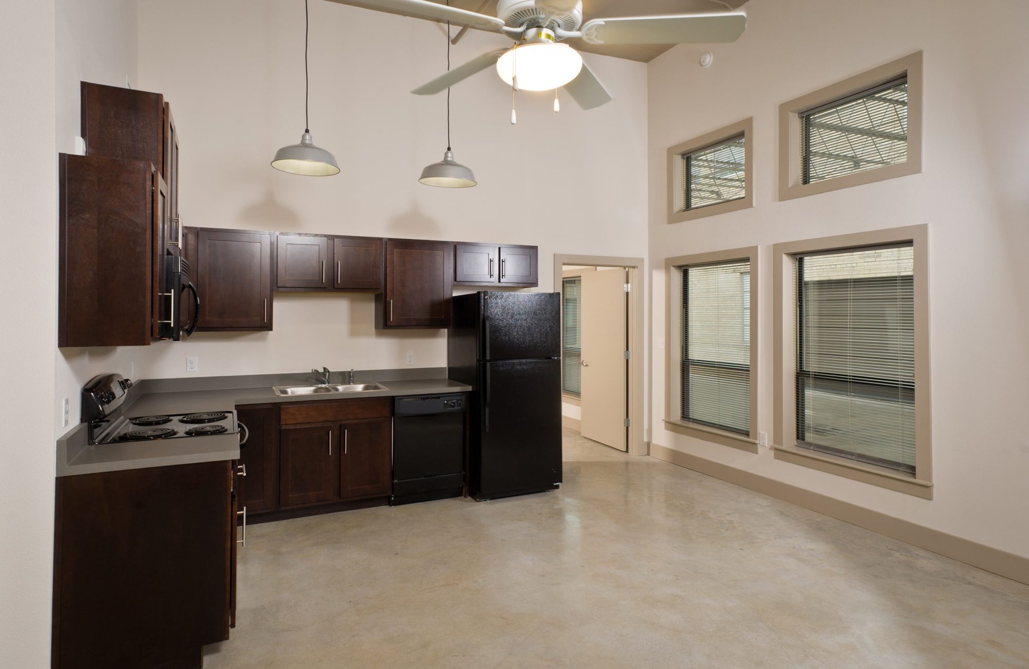 Modern apartment kitchen, unfurnished, with exposed industrial ceiling, large windows, espresso cabinets, and black appliances.