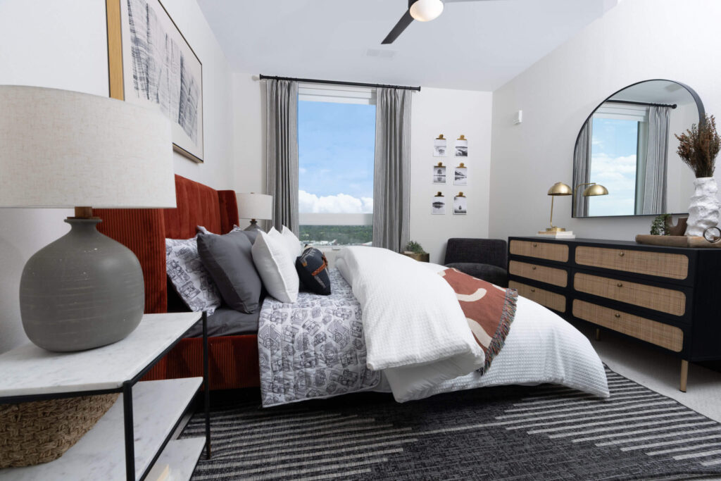Cozy bedroom with a red upholstered bed, layered pillows, modern dresser, and a large window with gray curtains.