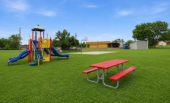 A red picnic table is in the foreground of a playground with a yellow and blue slide.