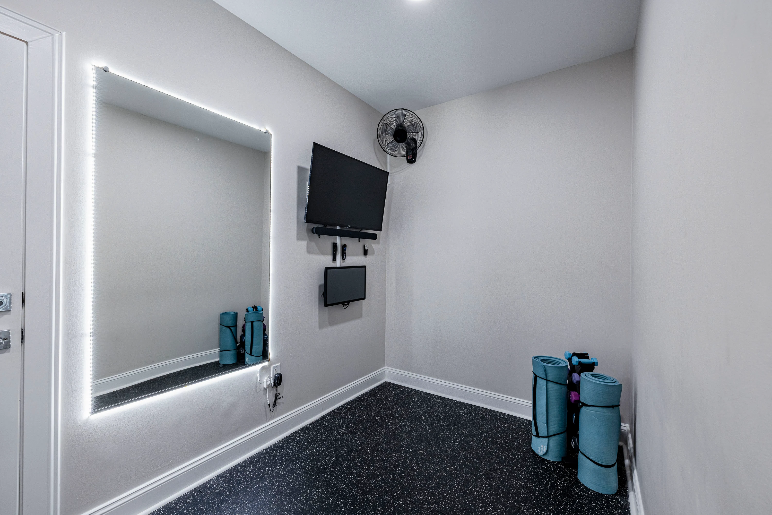 A modern apartment room featuring a large mirror, TV, fan, and exercise mats against a neutral wall.