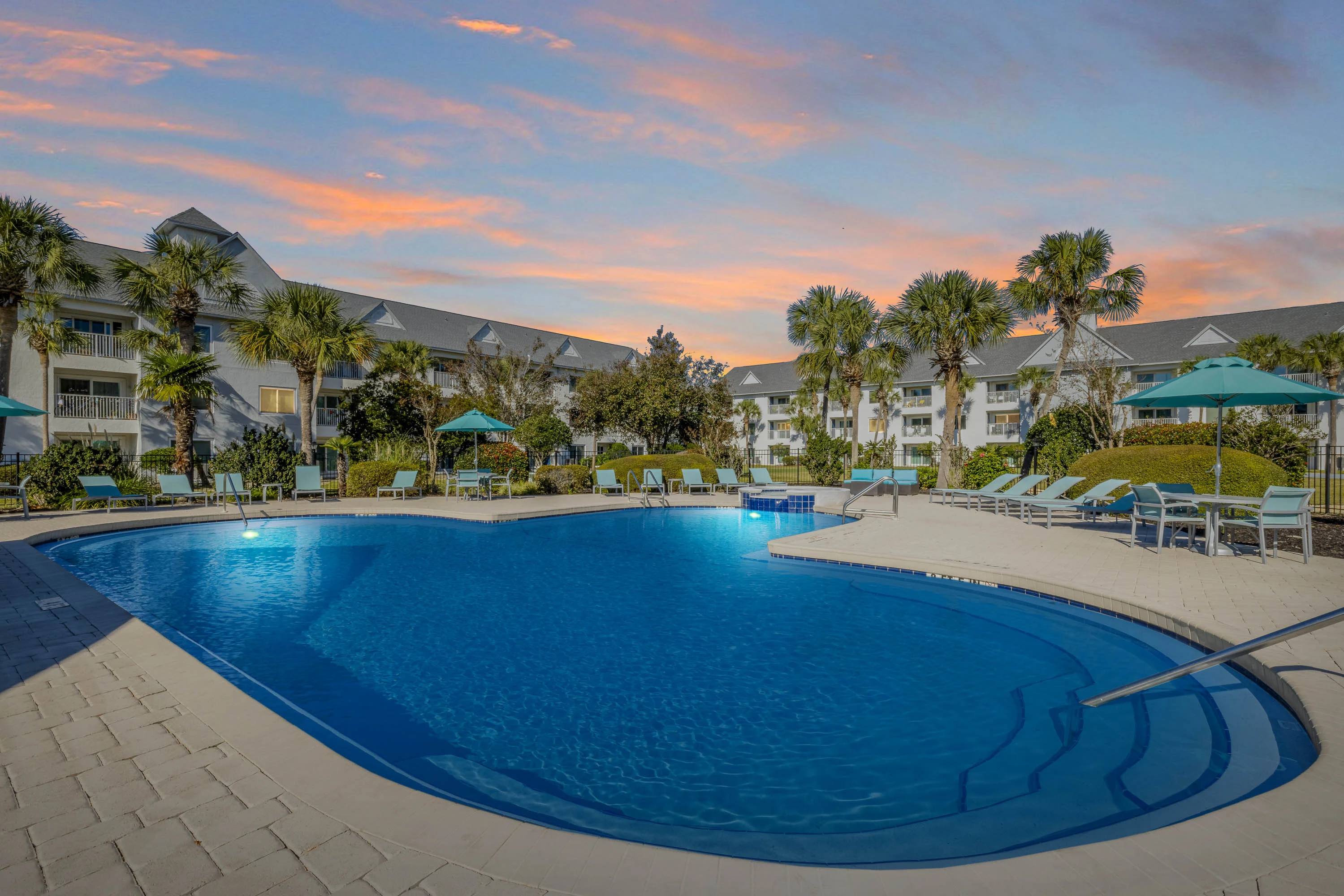 A serene apartment complex pool area surrounded by palm trees and lounge chairs, with a colorful sunset in the background.