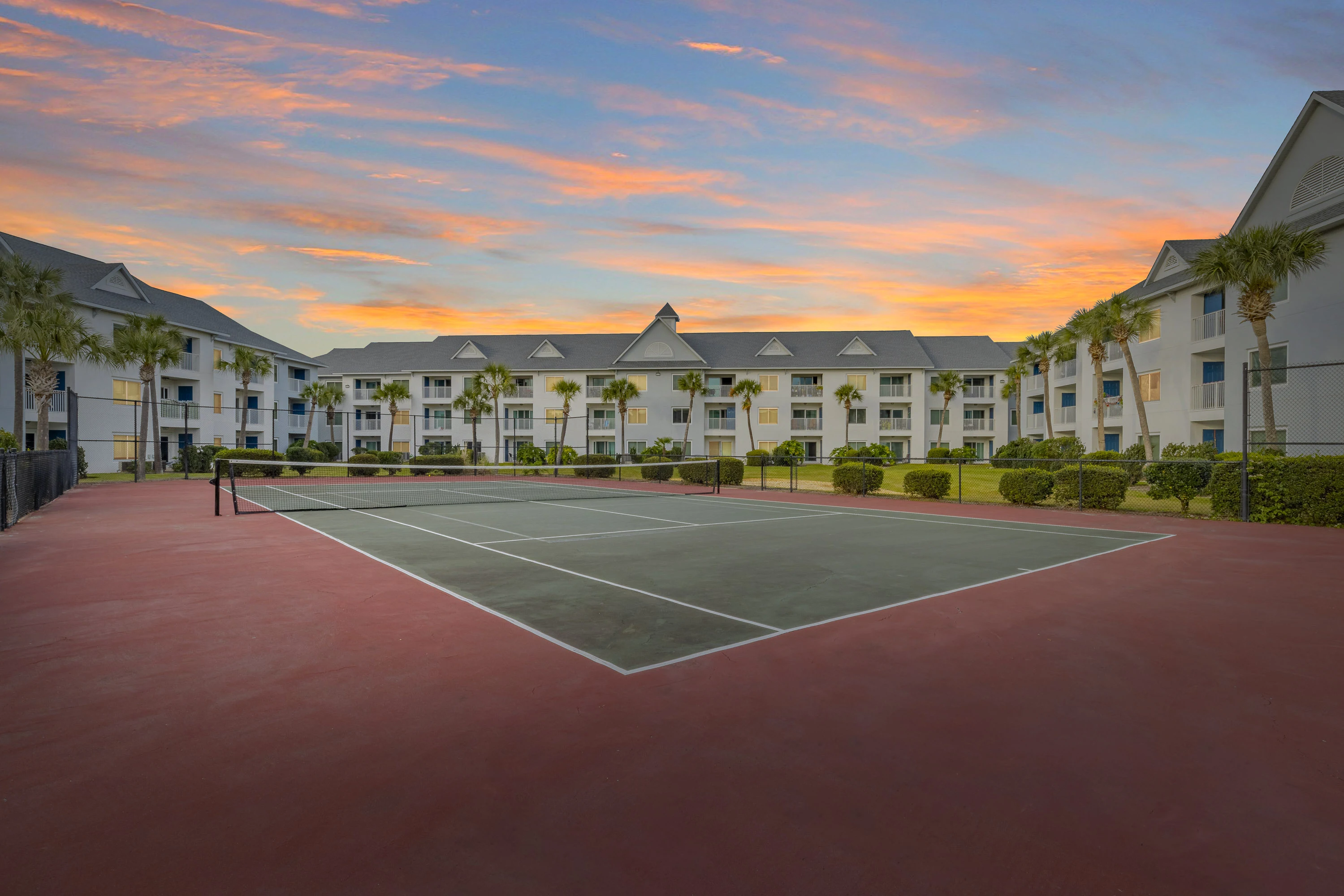 "View of an apartment complex with a tennis court in the foreground and a colorful sunset in the background."