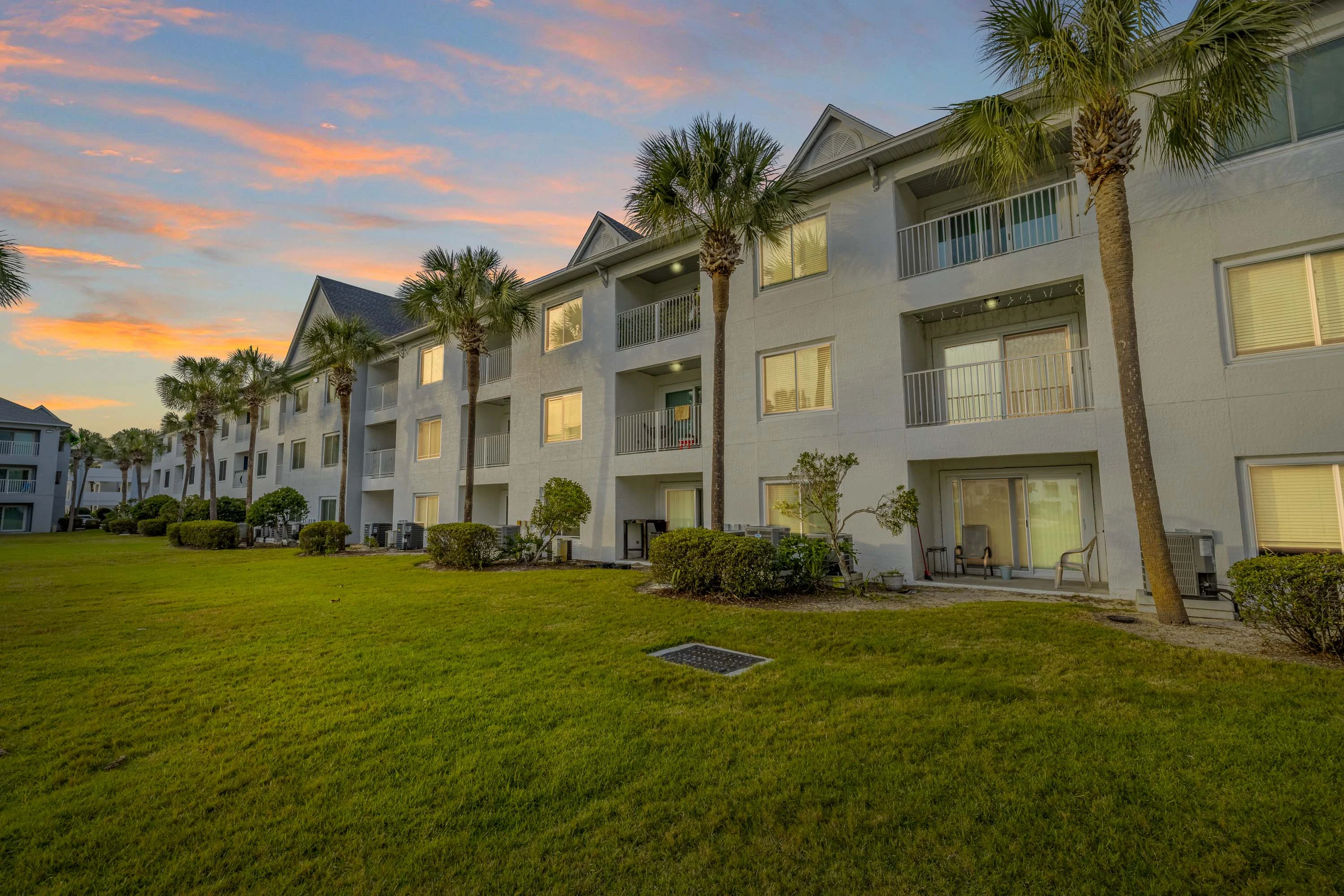 A peaceful apartment complex with palm trees, green lawns, and balconies, set against a colorful sunset sky.