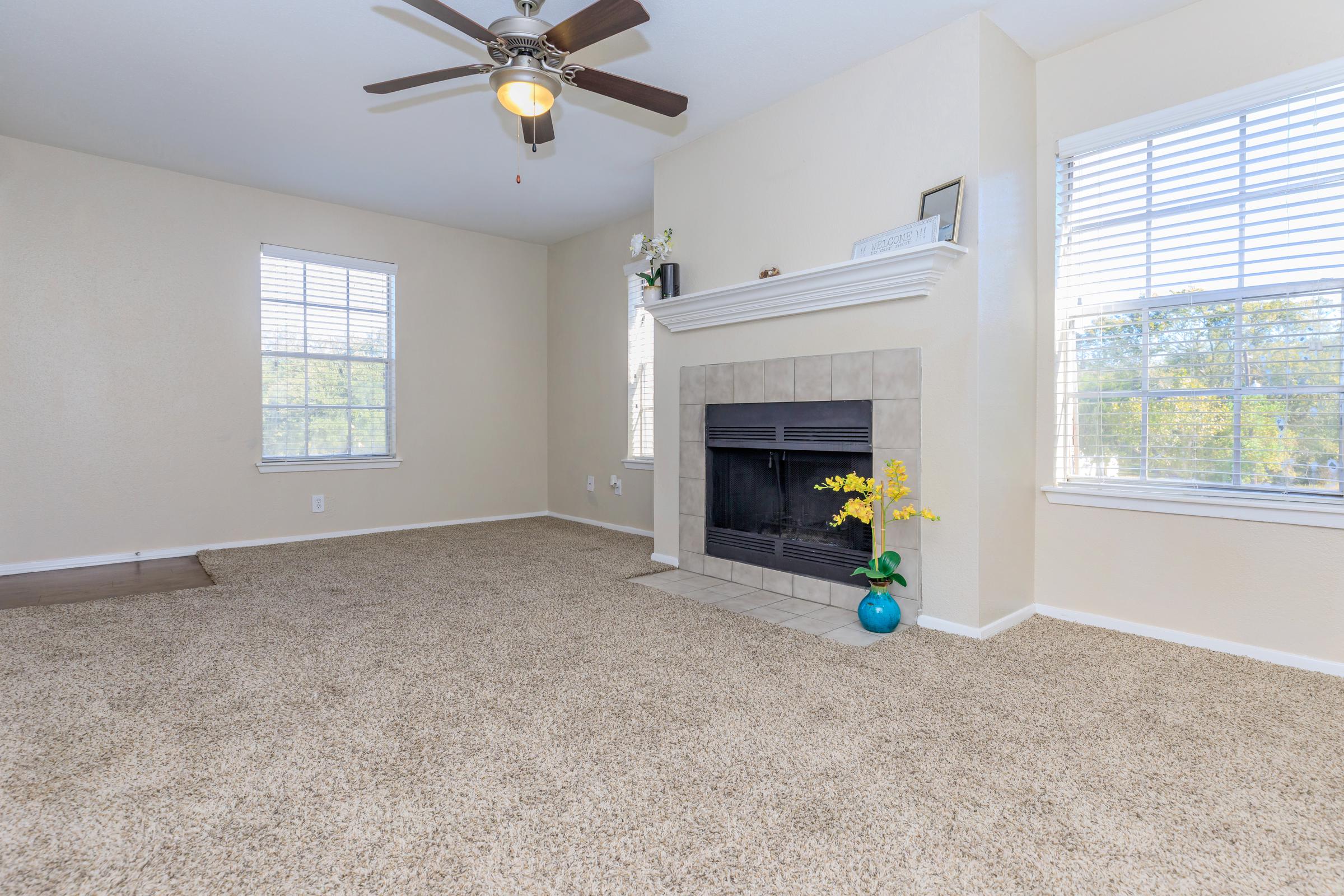 A spacious living room featuring a beige wall, a ceiling fan, and large windows that let in natural light. A tiled fireplace with a decorative mantel is centered on the wall. There is plush carpet on the floor and a potted yellow flower arrangement on the right side near the fireplace.