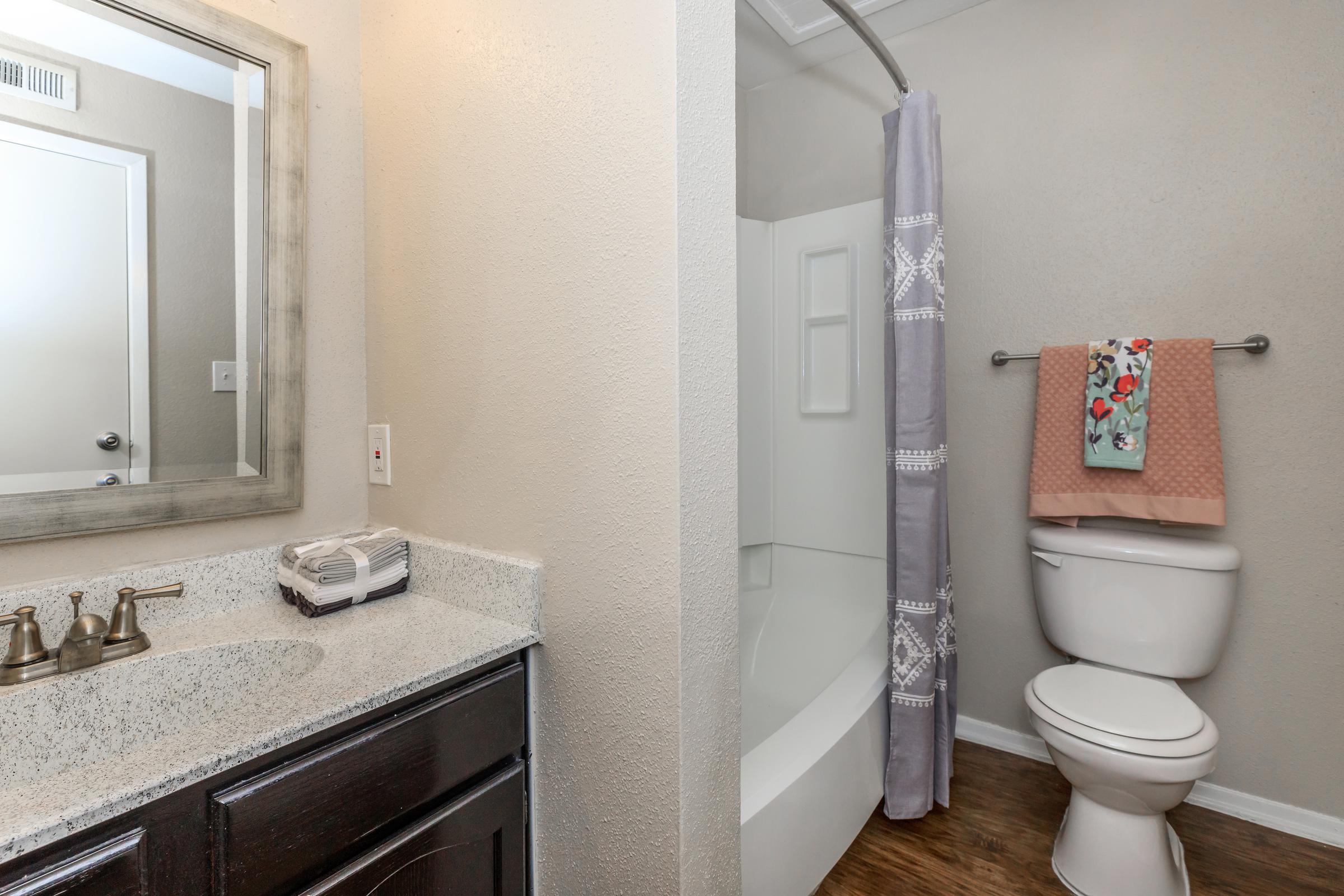 A clean bathroom featuring a modern vanity with a large mirror, granite countertop, and stylish faucet. To the right, a shower area with a gray curtain and a decorative towel on a rack. The walls are painted a light color, and the flooring is wooden, creating a warm and inviting atmosphere.
