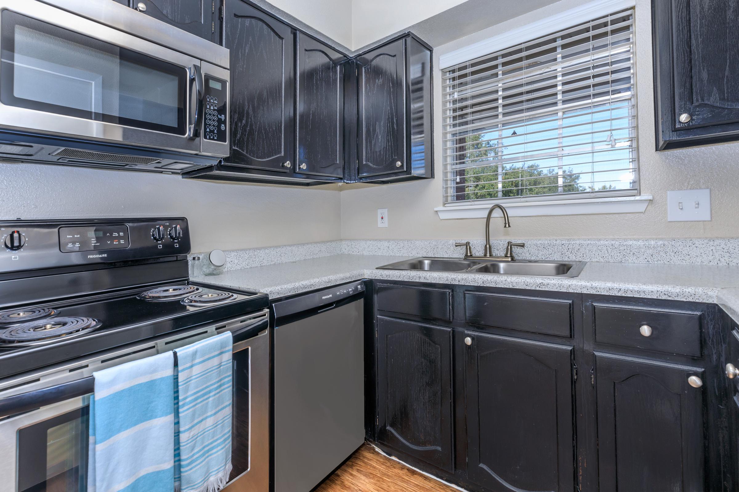 Modern kitchen featuring black cabinetry, a stainless steel microwave, an electric stove, and a dishwasher. A double sink is positioned beneath a window with blinds, allowing natural light. The countertop is gray with a textured finish, and a blue striped towel hangs from the stove. The floor has a wood-like appearance.