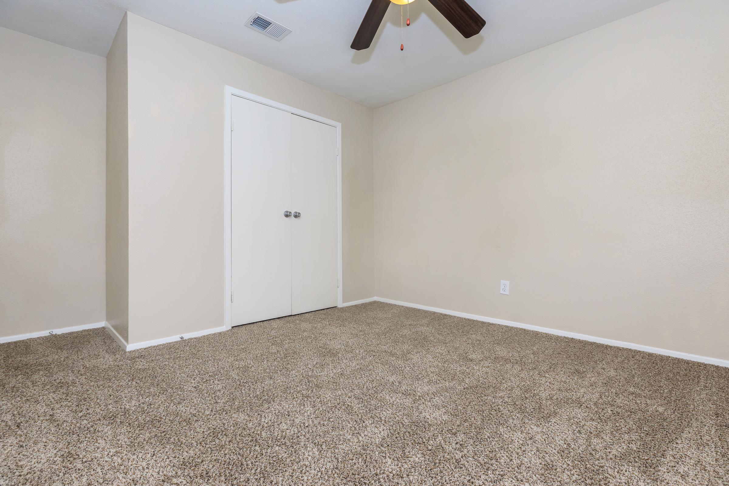 A vacant room with beige walls and carpet. A ceiling fan hangs from the ceiling. A closed white door is visible on one wall, while the space appears empty and well-lit, creating a neutral atmosphere. The flooring is covered in a light brown carpet.