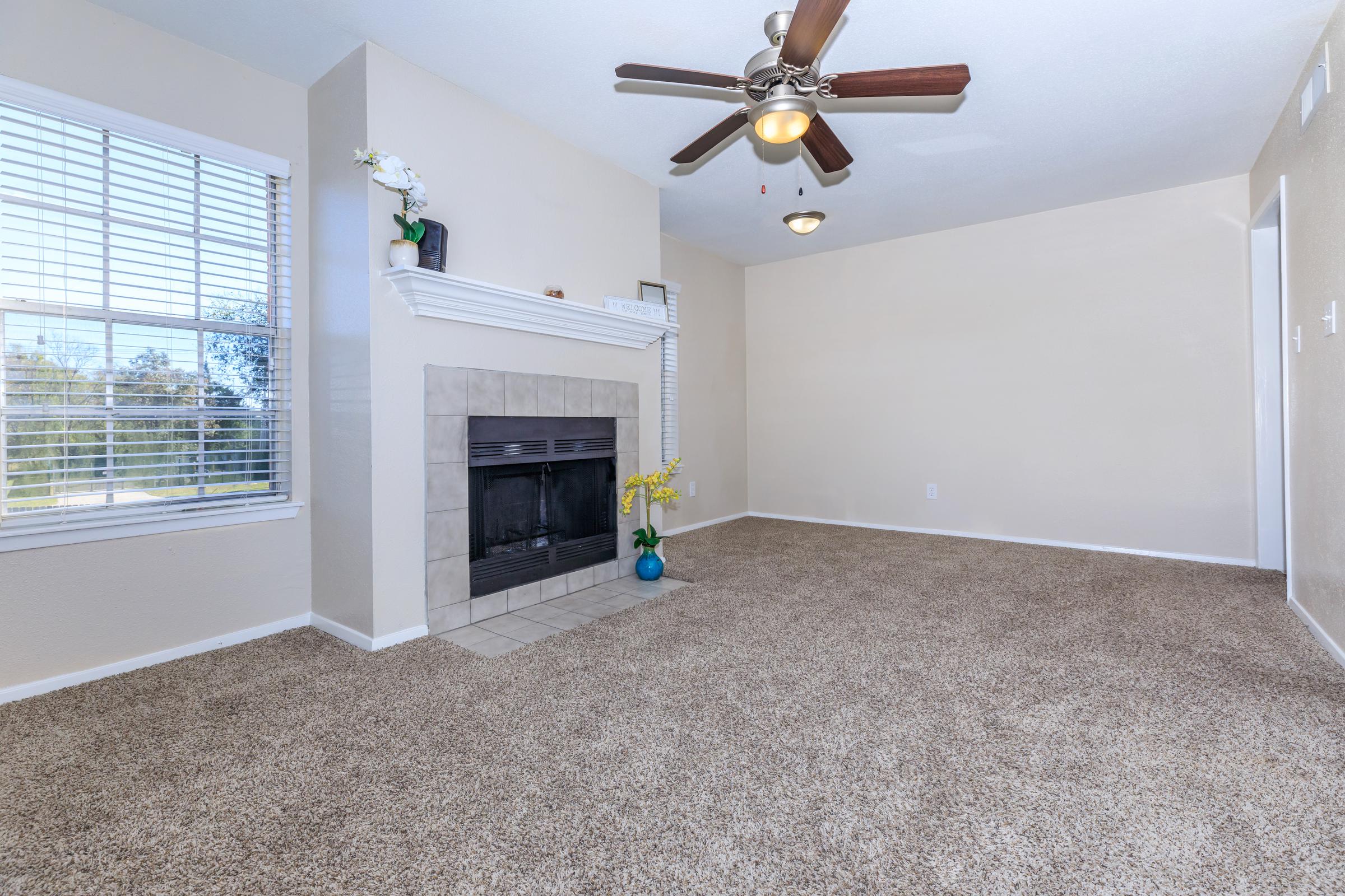 A spacious, well-lit living room featuring a beige wall color, a ceiling fan, and a large window with blinds. The area has plush, light brown carpet and a fireplace with a white mantel. A decorative plant is positioned on the mantel, enhancing the room's cozy atmosphere.