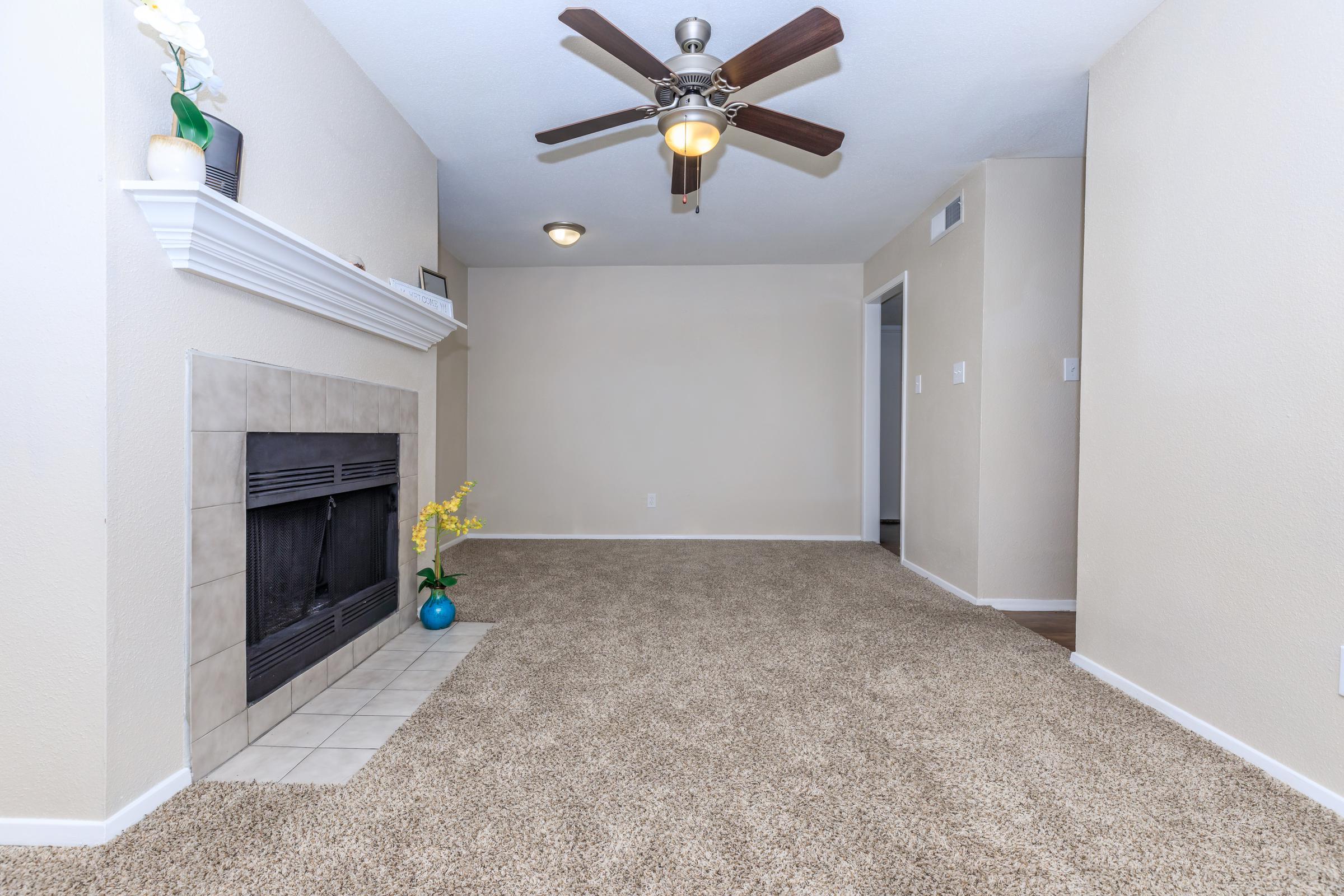 A spacious living room featuring a ceiling fan, carpeted floor, and a fireplace with a tile surround. Decor includes a potted plant on the hearth and a simple floral arrangement in a blue vase. The walls are painted light colors, enhancing the bright and airy atmosphere of the room.