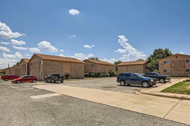Exterior view of a multi-building apartment community with parked cars and a blue sky.