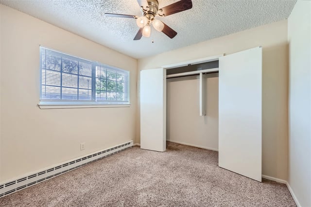 Bedroom with beige walls, carpet, window with blinds, and an open white closet with a hanging rod.