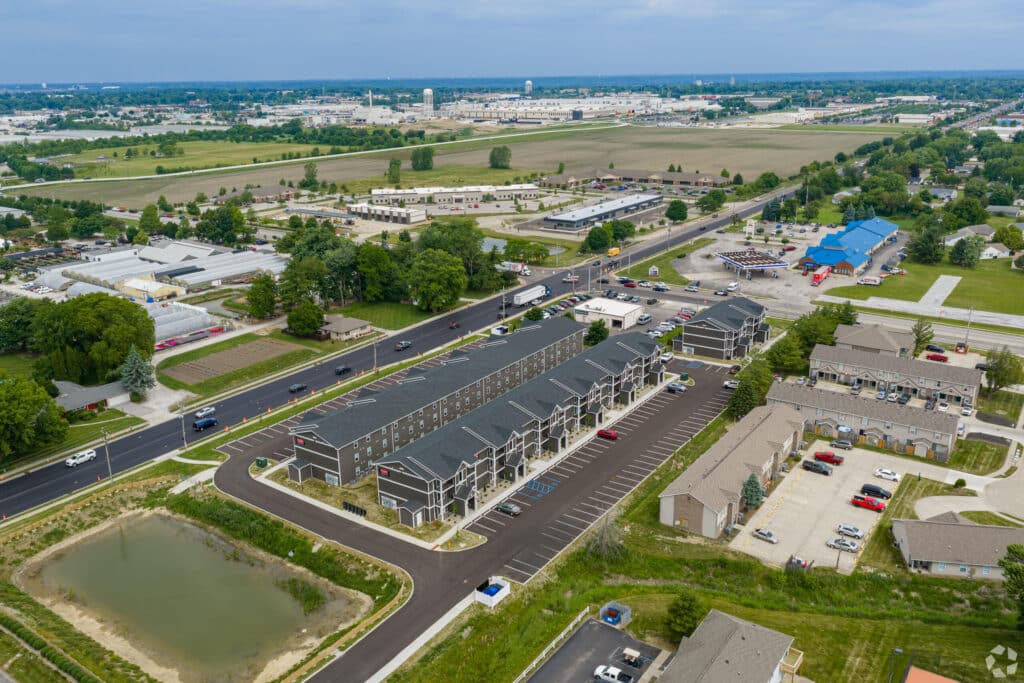 Aerial view of Gallery - Compass Flats, a residential townhouse complex with parking lots, nearby roads, and commercial buildings in a suburban area.