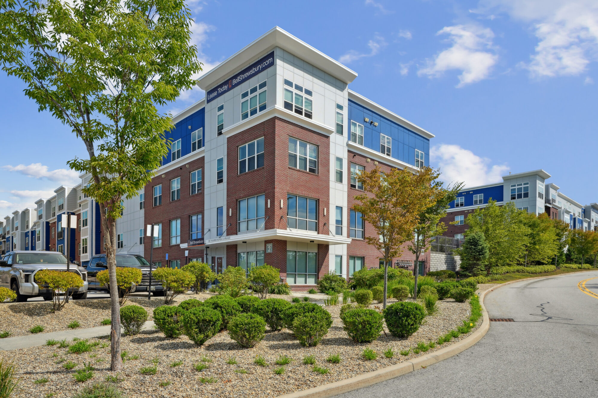Modern three-story apartment building in Littleton, CO with red brick and blue panel exterior, large windows, landscaped bushes and trees in front, and a curved road on a sunny day.