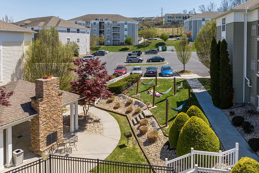 Charming apartment complex with landscaped gardens, cozy outdoor seating by a stone fireplace, and relaxing hammocks under sunny skies.
